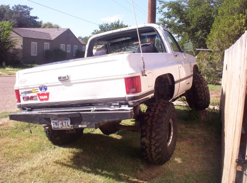 1988 Ford Bronco - This lifted GMC pickup truck is parked on uneven terrain next to a wooden fence.