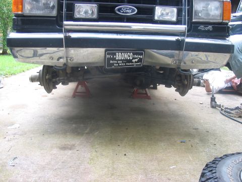1988 Ford Bronco - Front end of a Ford Bronco is elevated on jack stands, showing the suspension and a decorative bumper sticker.