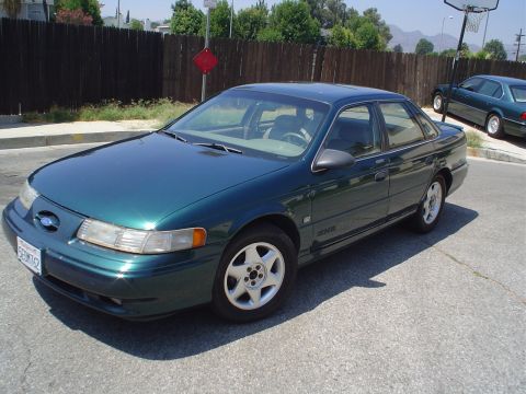 1993 Ford Taurus - This green sedan is parked on a street with a wooden fence and trees in the background.
