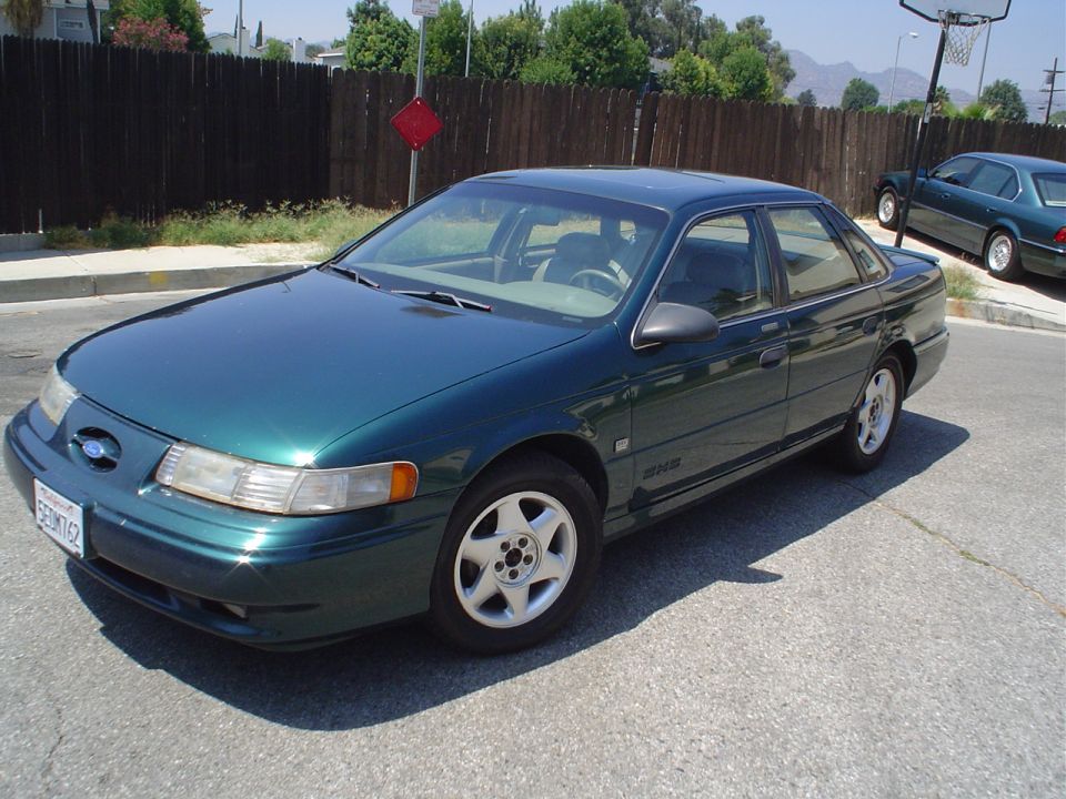 Green sedan parked on asphalt, another car in background. Basketball hoop visible