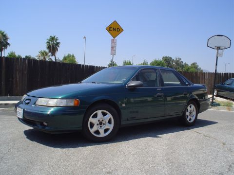 1993 Ford Taurus - A green sedan is parked on a paved surface with a basketball hoop and fence visible.