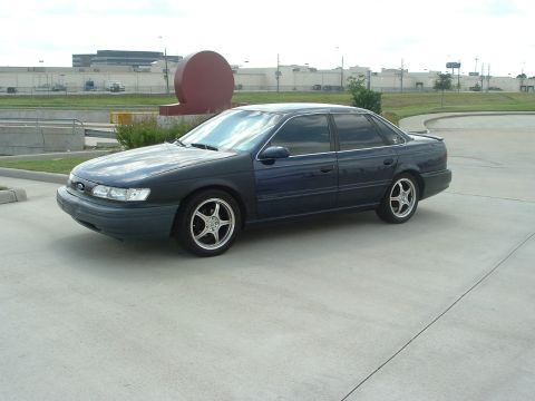 1993 Ford Taurus - Dark blue Ford Taurus sedan parked on a concrete lot, seen from the driver-side front three-quarter. It has polished aftermarket five-spoke…