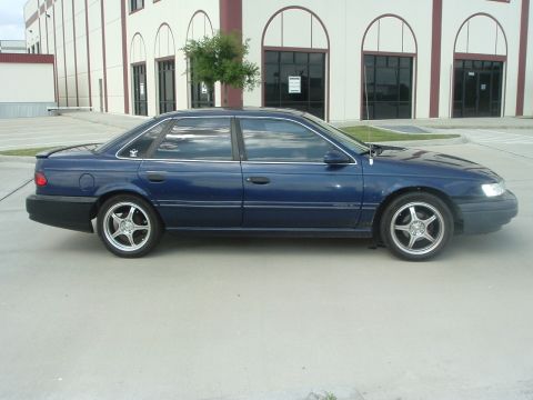 1993 Ford Taurus - Dark blue Ford Taurus four-door sedan photographed from the passenger side in a parking lot in front of a tan commercial building with arch…