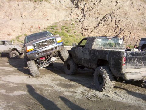 2000 Ford F-450 - Two modified trucks are navigating a rough, dirt terrain with a rocky hillside in the background.