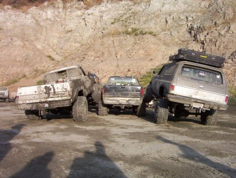 2000 Ford F-450 - Several modified trucks are parked off-road, covered in mud, with a rocky incline in the background.