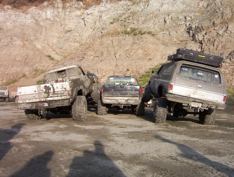 Three lifted, muddy trucks parked on dirt with rocky hillside behind. One SUV has a roof rack