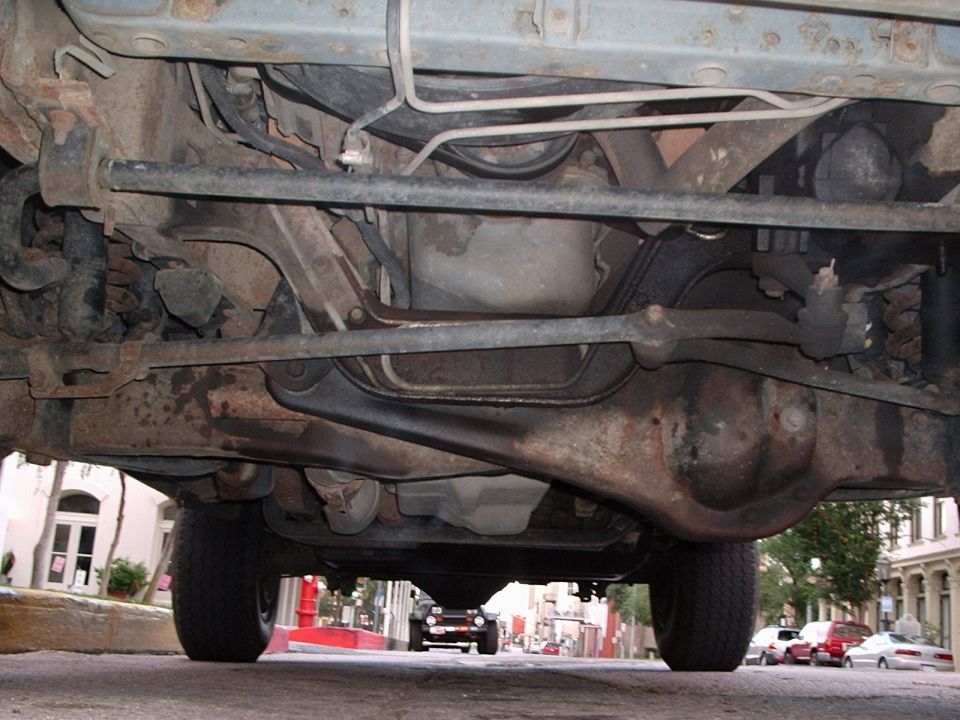 Underside of a vehicle with visible suspension and drivetrain components. A dark SUV is visible in the distance