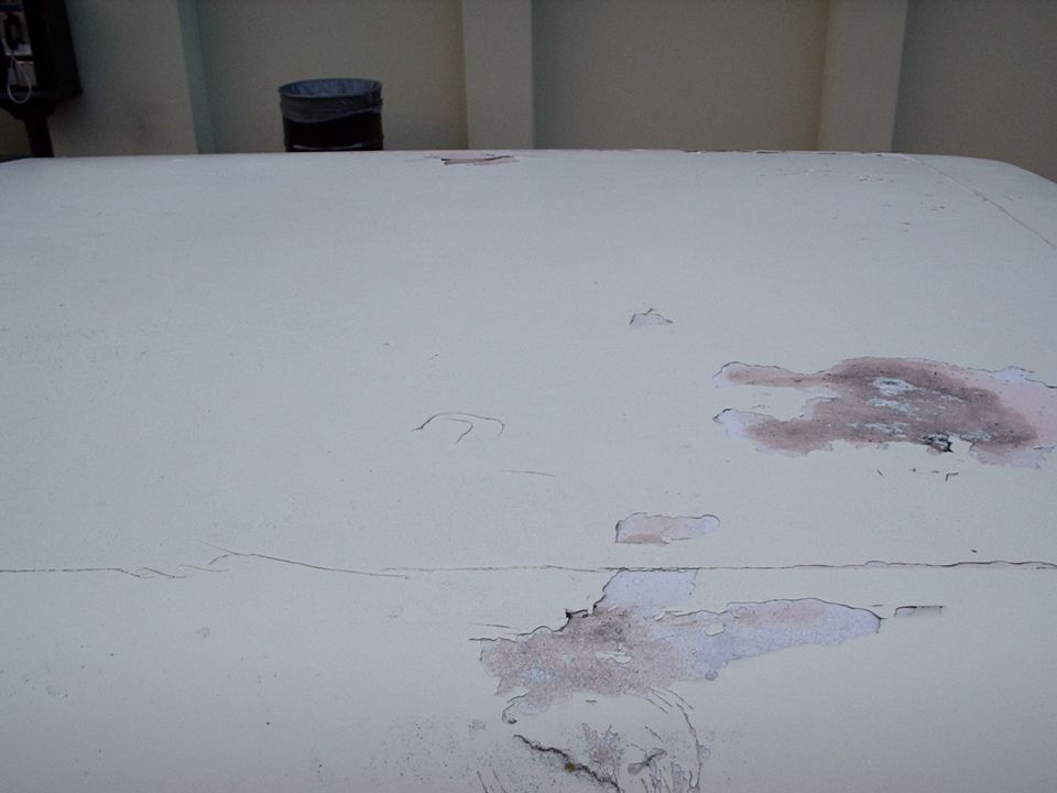Close-up of a white vehicle roof with peeling paint and rust spots. A trash can and part of a black object are visible in the background