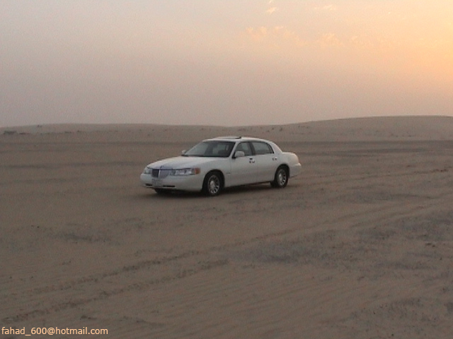 White sedan parked in a sandy desert with dunes under a hazy sky