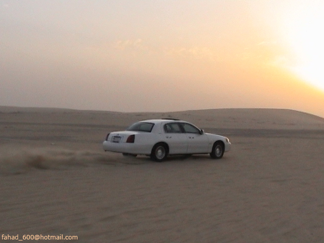 White sedan kicking up sand while driving across desert dunes at sunset