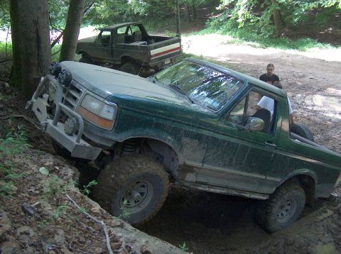 1989 Ford Bronco - Two Ford Broncos tackle a challenging off-road trail in a wooded area.