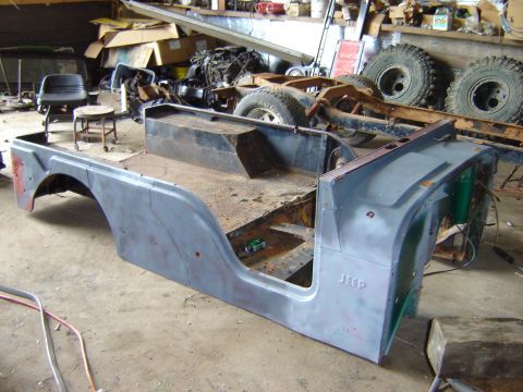 1992 Ford Bronco - The body tub of a Jeep CJ-6, shown in primer, sits in a workshop with other vehicle parts in the background.