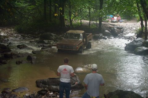 1987 Ford Bronco - A brown pickup truck navigates a shallow stream in a wooded area, with two onlookers observing from the shore.