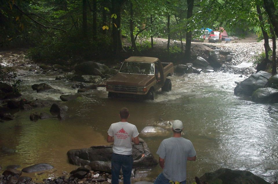 Brown pickup truck driving through a rocky stream, two men watching from the bank