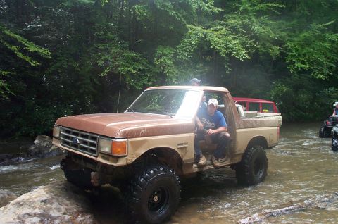1987 Ford Bronco - 1987 Ford Bronco navigates a rocky stream bed surrounded by lush green foliage.