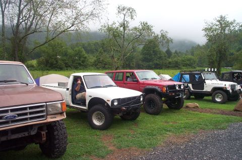 1987 Ford Bronco - A lineup of off-road vehicles is parked on a grassy area, with a mountainous landscape visible behind them.
