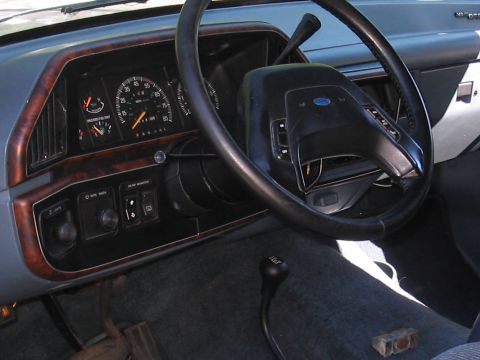 1979 Ford Bronco - Dashboard and steering wheel of a Ford truck, likely from the late 1980s, wood grain trim and a floor-mounted shifter.