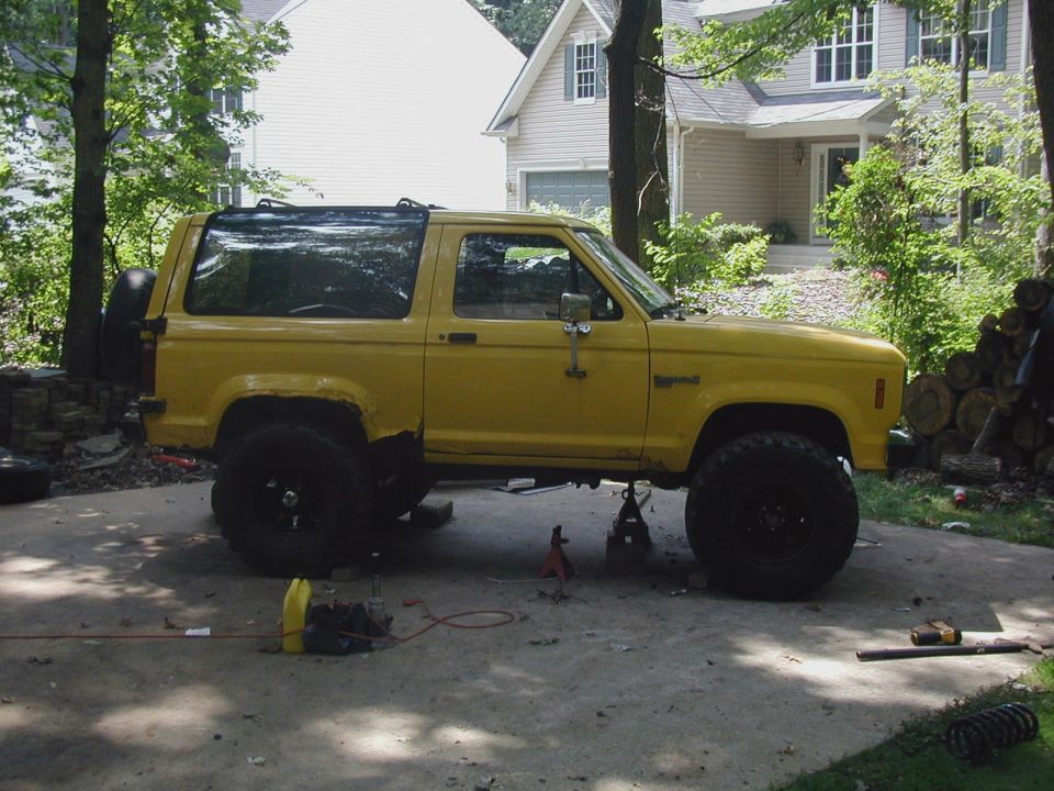 Yellow Ford Bronco II on jack stands in driveway, side view, with tools scattered around
