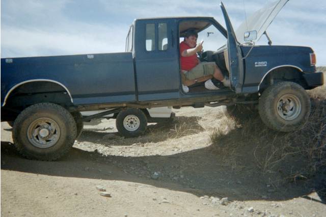 Person in red shirt sitting in a lifted dark Ford F-250 with door open off-road