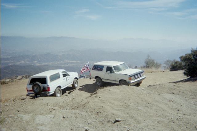 Two white Ford Broncos at a mountaintop overlook with US flag and hazy peaks