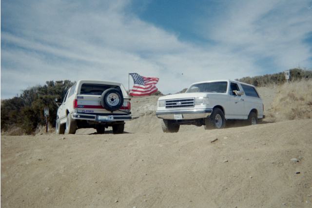 Two white Ford Broncos on a sandy hill with an American flag between them