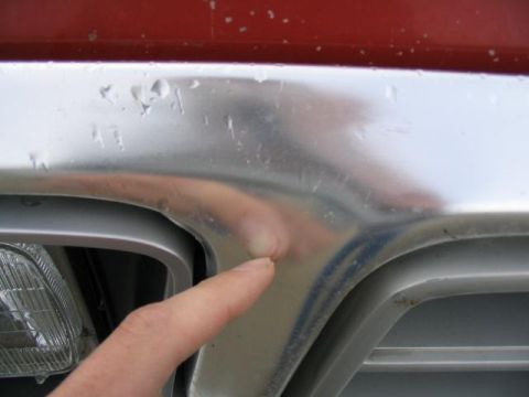 1979 Ford F-150 - A finger points to a dent on the chrome grille of a red truck. The grille shows signs of wear and damage.