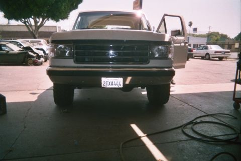 1990 Ford Bronco - Head-on view of a lifted tan Ford pickup with its driver door open, parked on pavement near other vehicles.