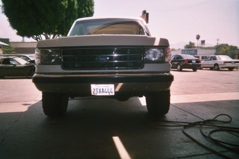 1990 Ford Bronco - Head-on view of a lifted Ford 4x4 pickup in a sunlit paved lot, with parked cars and a storefront behind.