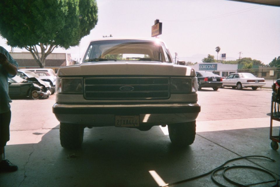 Front of a lifted tan Ford pickup in a lot with people and cars nearby