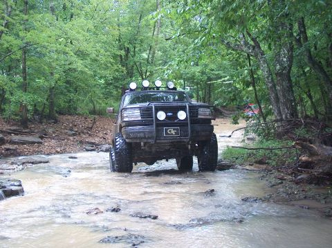 1989 Ford Bronco - This 1989 Ford Bronco navigates a muddy stream, equipped with auxiliary lights and a prominent GT emblem on its grille.