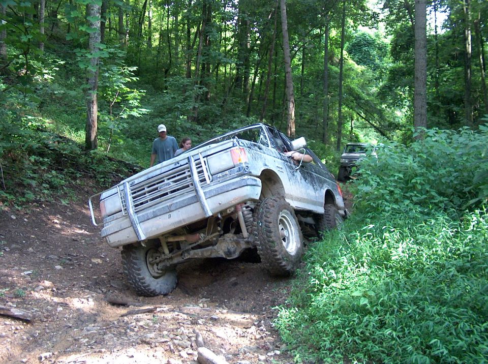 1989 Ford Bronco tilted on a muddy trail with another vehicle behind it in the woods