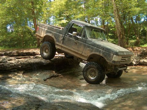 1989 Ford Bronco - A muddy Ford Bronco tackles an off-road stream crossing with one wheel lifted high.