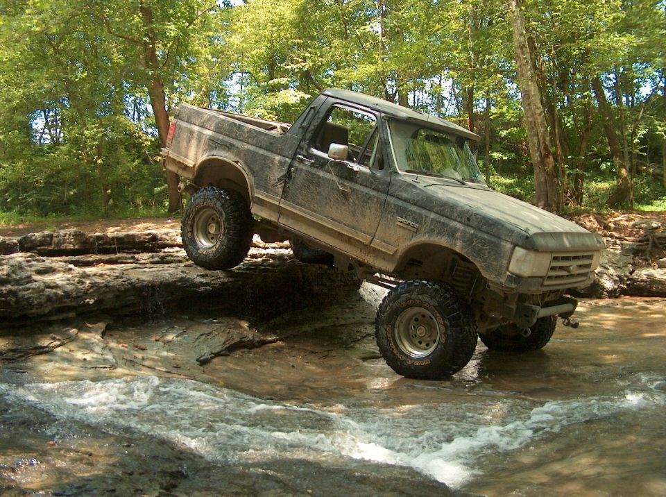 Mud-covered Ford Bronco navigating a rocky stream, front-right tire elevated