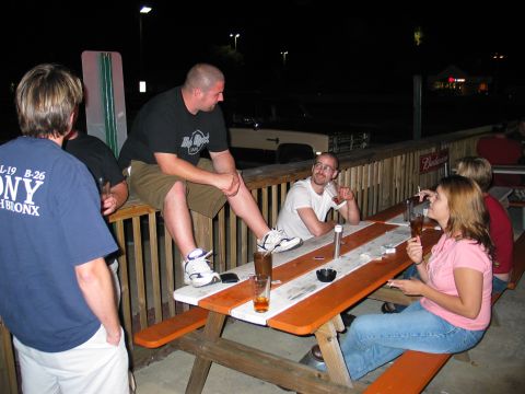 1995 Ford Bronco - Nighttime social scene on an outdoor bar patio, with people sitting and standing around orange-and-white picnic tables with beer glasses an…