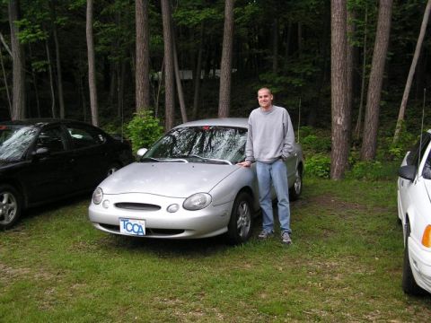 1998 Ford Taurus - A man poses with his silver 1998 Ford Taurus, parked outdoors with other vehicles nearby.