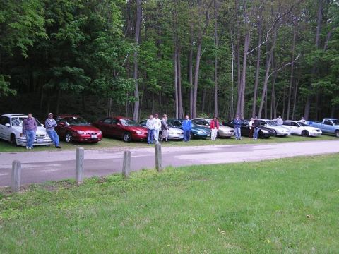 1998 Ford Taurus - Several cars are parked in a line next to a wooded area with people standing around them.