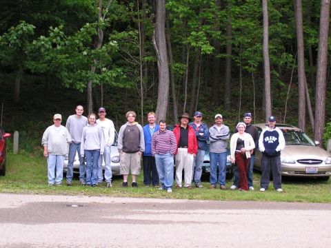1998 Ford Taurus - A group of people pose for a photo in front of several cars parked on a grassy area near a wooded background.