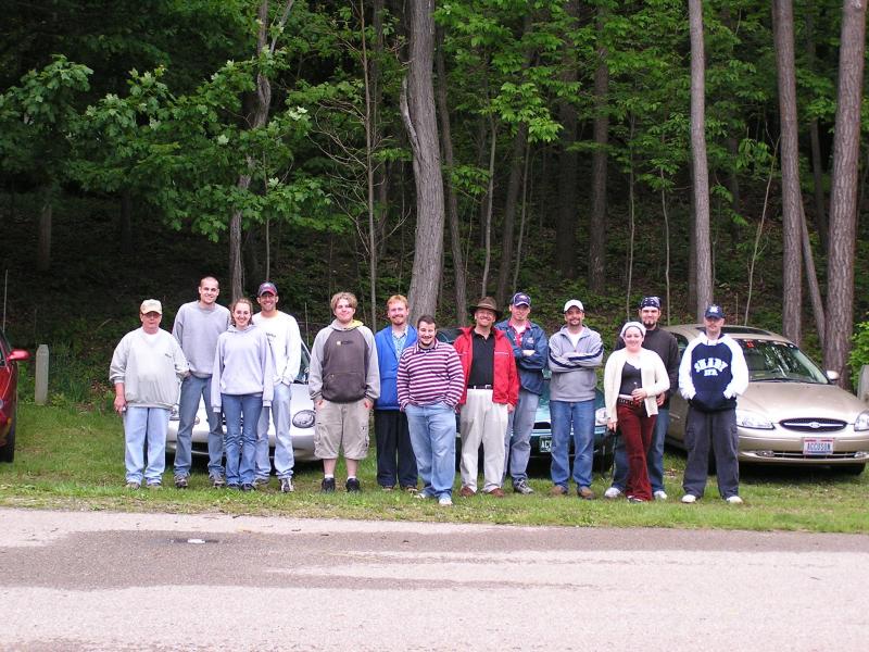 group of people standing in front of cars in a wooded area