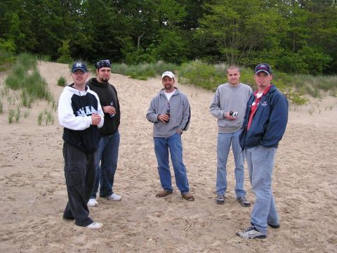 1998 Ford Taurus - A group of men gather on a sandy dune area, with trees lining the horizon. The image includes a SUPERMOTORS.NET watermark.