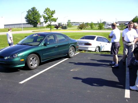 1998 Ford Taurus - Two sedans are parked in a lot on a sunny day, with a group of people observing.