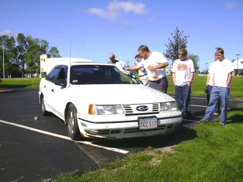 1998 Ford Taurus - A group of people are gathered around a white 1998 Ford Taurus, with one person cleaning the windshield.