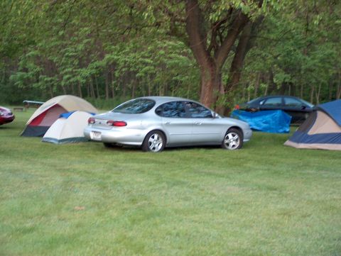 1998 Ford Taurus - A silver 1998 Ford Taurus is parked at a campsite, surrounded by tents and trees.