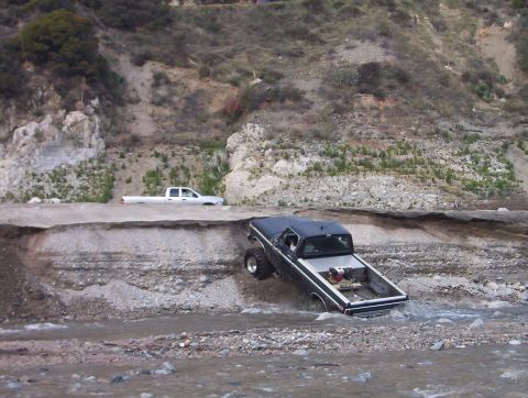 2000 Ford F-450 - A black square-body Ford pickup with white bedside stripe and mud tires descends a steep eroded bank into shallow water, its rear end alrea…