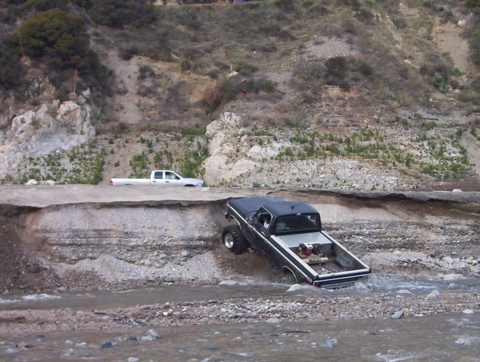 Black lifted Ford pickup dropping down an eroded bank into a shallow creek
