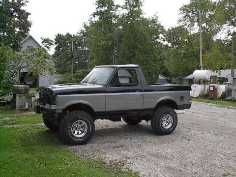 1989 Ford Bronco - This two-tone gray and black Ford Bronco is parked on a gravel lot with a barn and storage tanks in the background.