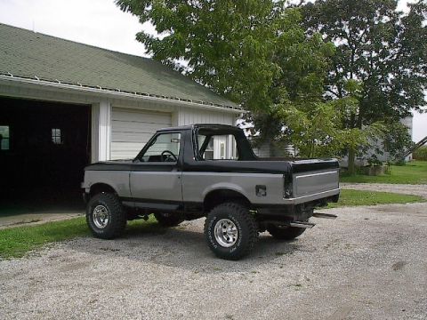 1989 Ford Bronco - This lifted Ford Bronco is parked outside a garage with a tree and rural buildings in the background.