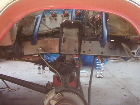 1978 Ford Bronco - In-progress front-end work seen through an orange-red fender opening, with the wheel off exposing the brake rotor, coil spring, blue-painte…