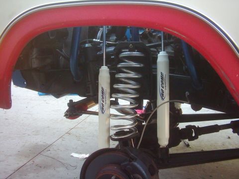 1978 Ford Bronco - View into the front wheel well of a lifted truck with the wheel removed, showing dual white Pro Comp shocks flanking a bare-metal coil spri…