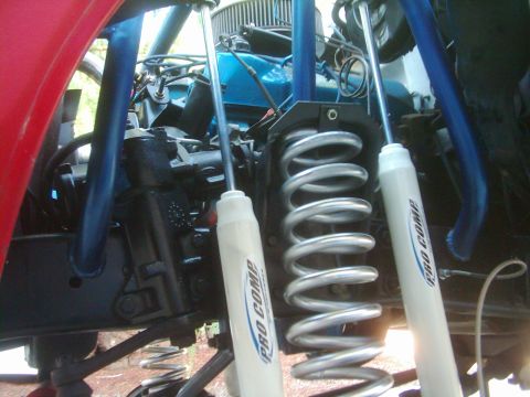 1978 Ford Bronco - Close-up looking up into the front suspension of a lifted truck, showing a bare silver coil spring flanked by two white Pro Comp shock abso…