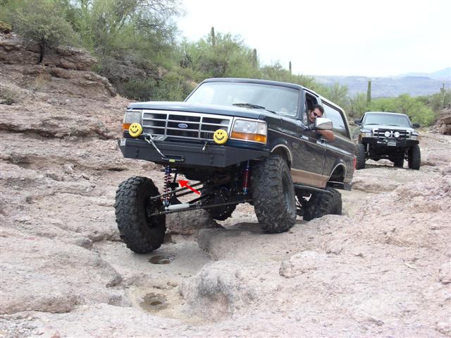 Lifted dark 1990s Ford Bronco with smiley light covers flexing its front axle on desert rock